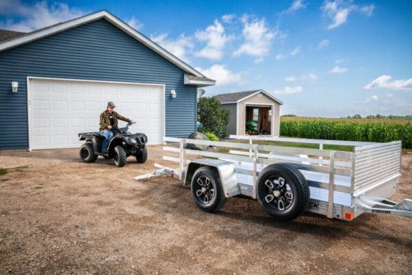 A single axle aluminum trailer with three side rail and a front rock guard being loaded by a guy on an atv in front of a blue garage with a white door and a cornfield with blue skies.