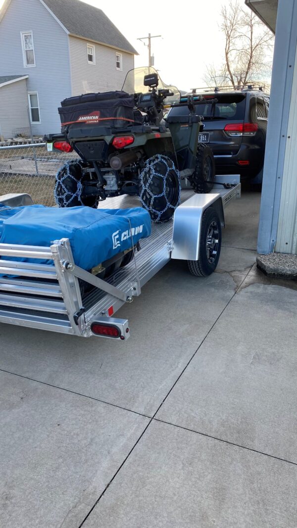 An all aluminum trailer with a bi-fold ramp loaded with a portable fish house and an ATV, pulled by a van.