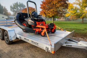 An orange and black lawn mower is secured to a trailer with ratchet straps. The trailer is parked in a driveway with autumn leaves on the ground.