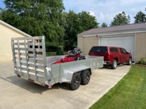An all aluminum trailer with side kits, front guard, and straight ramp, with a lawn mower on trailer, being pulled by a truck.