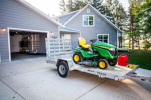 A green lawn mower is loaded onto a trailer on a driveway. The trailer has a straight ramp, wood deck, and aluminum frame. A house is in the background.