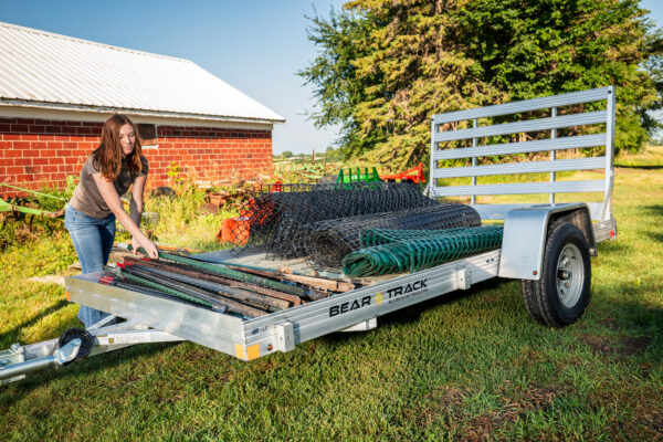 An aluminum framed trailer with a wood deck and a straight aluminum ramp being loaded with fencing posts by a girl.