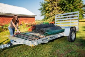 A girl is loading landscaping materials onto a trailer. The trailer has a straight ramp and an aluminum frame. There is a barn in the background.