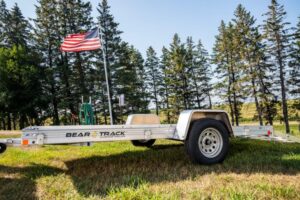 An aluminum framed utility trailer with a ramp down is parked in a grassy area with trees and a flag pole in the background.